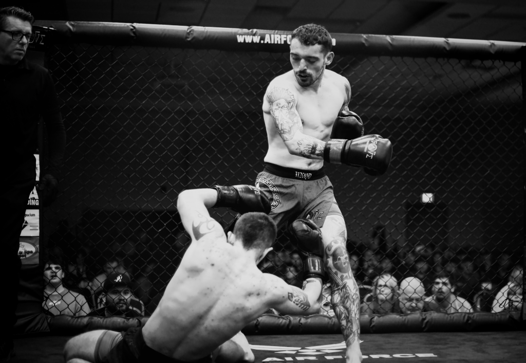 A black and white photograph captures an intense moment in a mixed martial arts (MMA) fight inside a cage. One fighter, standing, is delivering a powerful knee strike to his opponent, who is crouched on the ground. The standing fighter is shirtless, wearing shorts and gloves, with visible tattoos on his arms and legs. His expression is focused and determined. The grounded fighter, also shirtless, is attempting to defend himself, with his arms raised. A referee stands nearby, observing the action closely. The background shows a blurred crowd of spectators, adding to the atmosphere of the event. The cage's chain-link fence frames the scene, emphasizing the competitive nature of the sport.