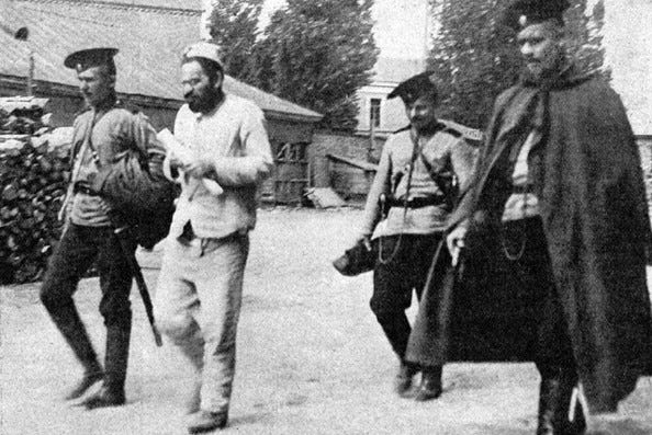 Black-and-white photo of a bearded man in light clothing walking between three uniformed guards with rifles, in what appears to be a courtyard with buildings and trees in the background.